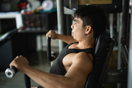 Asian man is working out at a gym, lifting weights. He is focused and determined, and his muscles are visible. Concept of strength and dedication to fitnessの写真素材