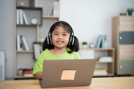 A young girl is sitting at a table with a laptop in front of her. She is wearing headphones and...