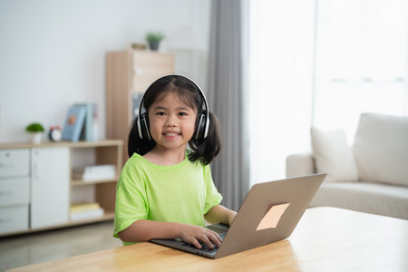 A young girl is sitting at a table with a laptop in front of her. She is wearing headphones and smiling. The room is filled with books and a clock on the wallの写真素材