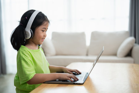 A young girl is sitting at a table with a laptop in front of her. She is wearing headphones and she is focused on her work. The room has a couch and a potted plant in the backgroundの写真素材