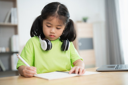 A young girl is sitting at a desk with a green shirt on and writing in a notebook. She is wearing headphones and she is focused on her workの写真素材