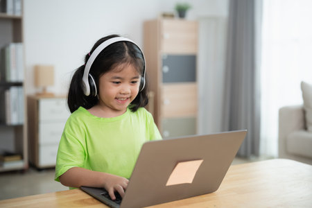 A young girl is sitting at a table with a laptop in front of her. She is wearing headphones and she is focused on her work. The room has a couch and a potted plant in the backgroundの写真素材