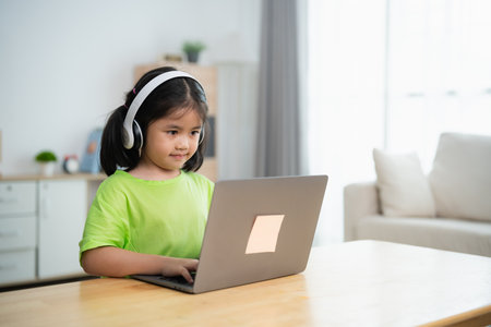 A young girl is sitting at a table with a laptop in front of her. She is wearing headphones and she is focused on her work. The room has a couch and a potted plant in the backgroundの写真素材