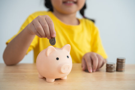 A young girl is sitting at a table with a piggy bank and a pile of coins. She is putting coins into the piggy bank, which suggests that she is saving moneyの写真素材