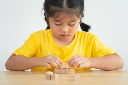 Asian young girl is sitting at a table with wooden blocks in front of her. She is focused on building a structure with the blocksの写真素材