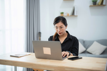 Asian woman is sitting at a desk with a laptop and a cell phone. She is wearing glasses and she is focused on her work. The room has a couch and a few books scattered aroundの写真素材
