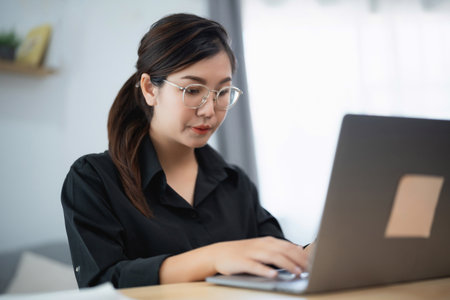 Asian woman wearing glasses is typing on a laptop. She is focused on her work and she is in a serious moodの写真素材
