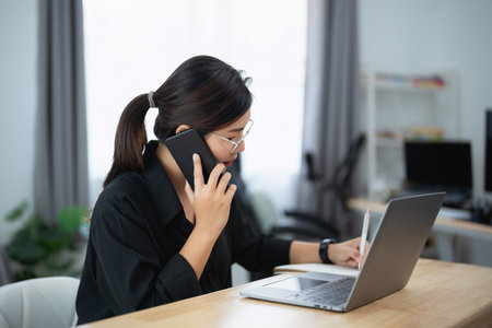 Asian woman is talking on her cell phone while sitting at a desk with a laptop. She is wearing glasses and she is focused on her conversation. The scene suggests a busy workday or a moment of multitaskingの写真素材