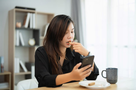 Asian woman is eating a snack while looking at her cell phone. She is sitting at a table with a cup and a plate of foodの写真素材