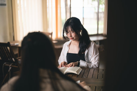 A woman is reading a book at a table. She is wearing glasses and has a ponytail. Another woman is sitting at the table next to herの写真素材