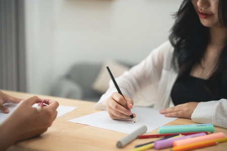 A woman is writing with a pencil on a piece of paper. She is sitting at a table with a variety of colorful markers and crayons. The scene suggests a creative and artistic atmosphereの写真素材