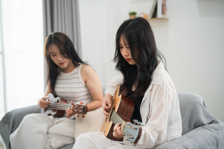 Two women are playing guitars on a couch. One of them is wearing a white shirt. Scene is relaxed and casualの写真素材