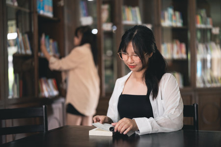A woman is sitting at a table in a library, reading a book. She is wearing glasses and a black shirtの写真素材