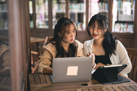 Two women sitting at a table looking at a laptop. One of them is wearing glasses. Scene is friendly and collaborativeの写真素材