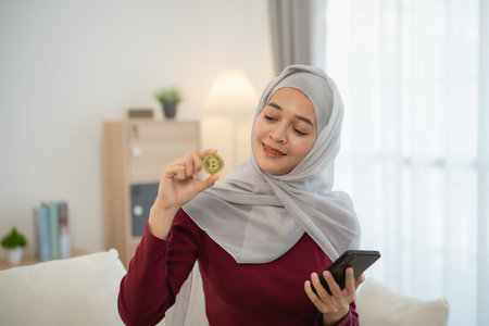 Muslim islam woman with hijab wearing a grey scarf is holding a phone and a yellow coin. She is smiling and she is happyの写真素材