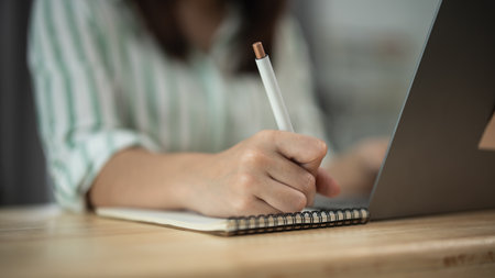 Asian woman is writing in a notebook with a pen. She is sitting at a desk with a laptop in front of her. The scene suggests that she is working or studying, possibly taking notes or writing a reportの写真素材