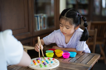 Asian young girl is playing with a game that has a lot of colorful balls. She is using chopsticks to pick up the balls. A woman is watching herの写真素材