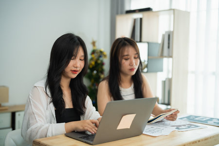 Two Asian women are sitting at a table with a laptop and a stack of papers. They are working together on a projectの写真素材