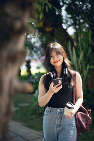 A cheerful young woman enjoys a sunny day in a park, casually dressed with headphones and holding her smartphone and coffee, exuding relaxation and happiness.の写真素材