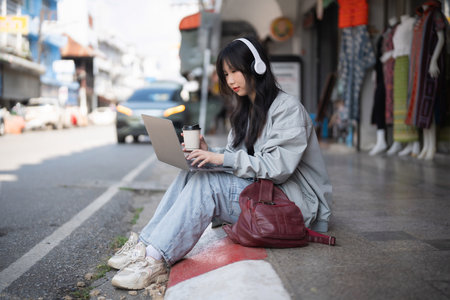 A young woman sits on a street curb, using her laptop while sipping coffee. She wears headphones and casual attire, embodying a modern and creative lifestyle.の写真素材