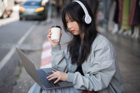 A young woman sits outdoors with her laptop and a cup of coffee, wearing headphones as she focuses on her work in a vibrant urban setting.の写真素材