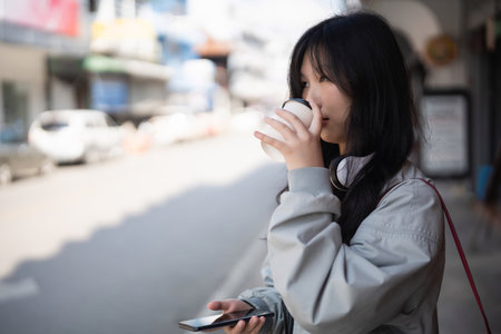 A young woman in a casual jacket enjoys sipping coffee while standing on a street. She casually holds her smartphone, embodying a modern urban lifestyle on a sunny day.の写真素材