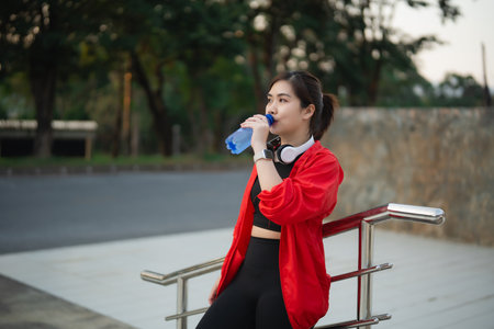A young woman in bright sportswear sipping water while sitting outdoors. She embodies a healthy lifestyle, balancing fitness with relaxation in a beautiful natural setting.の写真素材