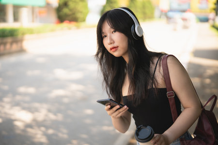 Young Asian woman with headphones enjoying music while holding smartphone and coffee cup in urban outdoor setting on a sunny dayの写真素材