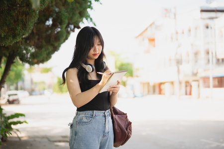 Young Asian woman with headphones writing notes outdoors in a sunny urban setting, reflecting creativity and inspiration among city life sceneryの写真素材