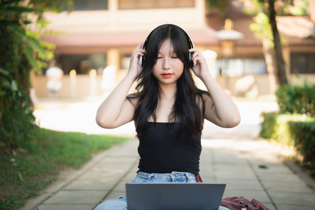 Young Asian Woman Wearing Headphones Enjoying Music While Working on Laptop in Outdoor Park Setting Surrounded by Greenery and Natural Lightの写真素材