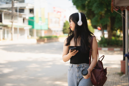 Young Asian woman in casual outfit with headphones holding smartphone and coffee walking in tranquil urban street on sunny day surrounded by greeneryの写真素材