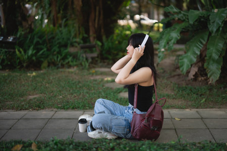 Young Asian woman with headphones enjoying music in green park, sitting on a path surrounded by plants, casual fashion and coffee cup beside herの写真素材