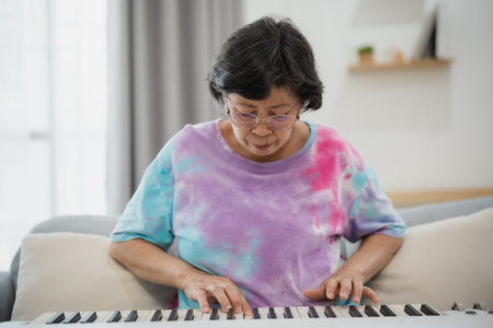 Senior Asian woman enjoying music while playing keyboard at home, wearing comfortable clothes, practicing melody in cozy living room settingの写真素材