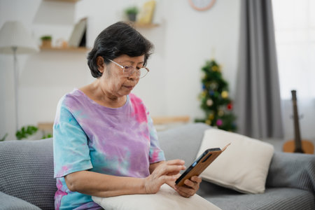 Elderly Asian woman with glasses reading smartphone while sitting on couch at home in cozy and bright living room with curtains and soft pillowsの写真素材