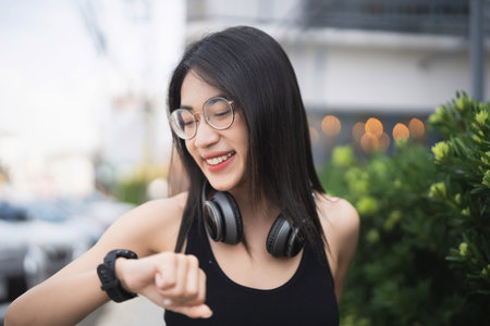 A cheerful young woman is outdoors, wearing wireless headphones and checking her smartwatch. The...