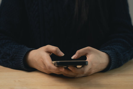 A close-up view of a person's hands holding a smartphone, showcasing intimate interaction with technology while sitting comfortably at a wooden table.の写真素材