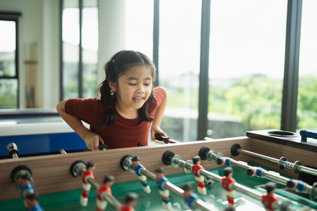Joyful Girl Enjoys Foosball Game in Bright Room with Large Windows Overlooking Green Outdoors,...