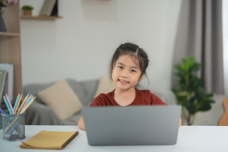 Smiling young girl using laptop at home for online learning, study, and education in cozy living room with natural light and comfortable atmosphereの写真素材