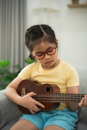 Young girl with glasses playing ukulele while sitting comfortably in living room, showcasing talent and joy of music in home environmentの写真素材