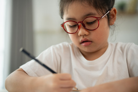 Young Girl with Glasses Focused on Writing in Notepad, Capturing The Essence of Creativity and Learning in a Bright Indoor Environmentの写真素材