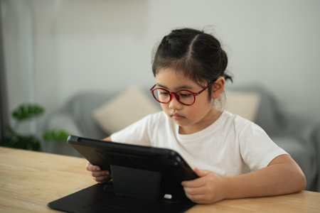 Young girl wearing glasses, focused on tablet screen while sitting at a wooden table in a bright, modern indoor setting with soft furnishingsの写真素材