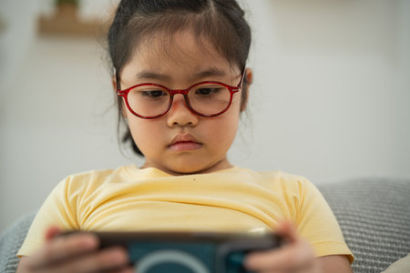 Young girl with red glasses focused on smartphone, engaging with digital content, enjoying technology in cozy indoor setting, playful mood, casual wearの写真素材