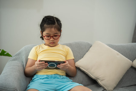 Focused Young Girl Engaged in Interactive Game on Smartphone While Sitting Comfortably on Soft Couch Surrounded by Bright Indoor Spaceの写真素材