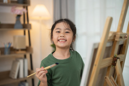 Cheerful Young Girl Smiling While Holding a Paintbrush in Front of an Easel in a Bright Indoor Art Space with Natural Lightの写真素材