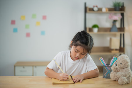 Young girl with black hair writing on paper at a wooden table in a cozy indoor environment surrounded by toys and colorful post-it notes on the wallの写真素材