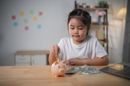 Young girl happily saving coins in a piggy bank with cash on the table in a cozy indoor environment, promoting financial literacy and smart habitsの写真素材