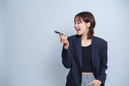 Young woman in oversized black blazer laughing while talking into voice recorder on light blue...