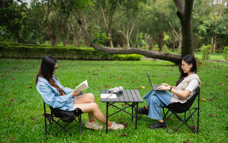 Two Young Women Engaged in Leisure Activities Outdoors in a Lush Green Park, One Reading a Book...