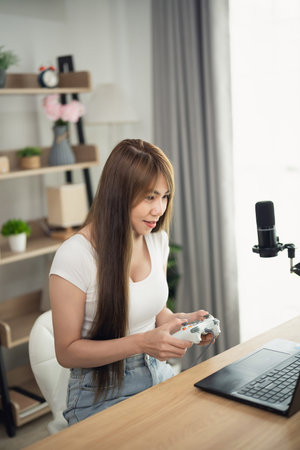 Young woman playing video games at home, streaming on a laptop with microphone, engaged in...