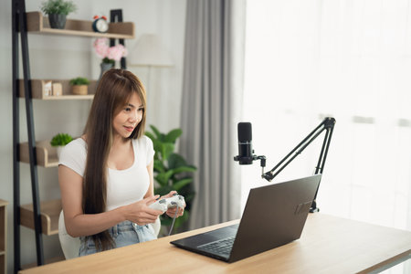 Young woman enjoying gaming session at home, playing video games while using laptop and microphone...